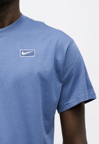 Άτομο που φοράει ένα απλό μπλε t-shirt Nike με κεντητό λευκό λογότυπο swoosh στο στήθος, πάνω σε απλό φωτεινό φόντο.