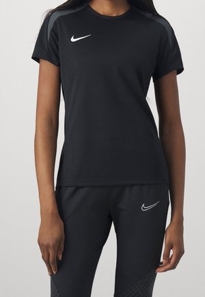 Kobieta w czarnej sportowej koszulce Nike z krótkim rękawem i czarnych spodniach z białymi logo Nike, stojąca na jednolitym tle.