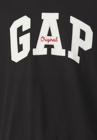 Camiseta de algodón negra con grandes letras blancas "GAP" y un acento rojo "Original" abajo. Diseño simple y clásico con una textura suave.