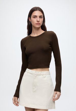 Mladá žena s dlhými hnedými vlasmi oblečená v tmavohnedom dlhom rukáve crop top a svetlo béžovej sukni na jednoduchom pozadí.