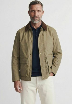 Homme à la barbe grise portant une veste olive avec un col marron sur une chemise bleu marine et un pantalon blanc, debout devant un fond uni.