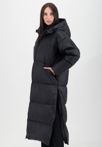 Doudoune noire avec capuche, au design long, texture matelassée, poches latérales et accent de marque le long de l'ourlet.