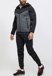 Survêtement de sport noir et gris comprenant une veste à capuche zippée avec un design sur le panneau avant, associée à un pantalon fuselé assorti et des baskets.