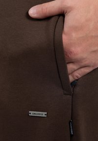 Hand teilweise in der Tasche von braunen Stoffhosen, mit einem kleinen Metalletikett mit der Aufschrift "SMILODOX."