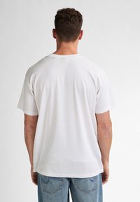 Tricou alb din bumbac cu mâneci scurte, croială relaxată și decolteu rotund. Textură netedă, fără modele, vedere din spate cu blugi din denim albastru.