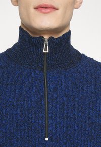 Gestrickter Pullover in Dunkelblau mit strukturiertem Muster und hohem Kragen. Der Reißverschluss an der Vorderseite mit metalischer Hardware reicht bis zum Kragen.