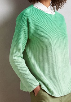 Personne portant un pull en maille côtelée vert clair par-dessus une chemise blanche à col, une main dans la poche d'un pantalon vert olive.