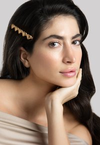 Goldener herzförmiger Haarclip schmückt dunkles, welliges Haar. Das Modell hat glatte Haut, trägt ein hellbeiges schulterfreies Oberteil, vor neutralem Hintergrund.