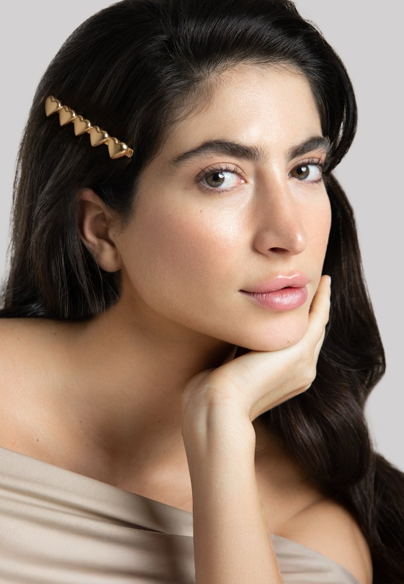 Goldener herzförmiger Haarclip schmückt dunkles, welliges Haar. Das Modell hat glatte Haut, trägt ein hellbeiges schulterfreies Oberteil, vor neutralem Hintergrund.