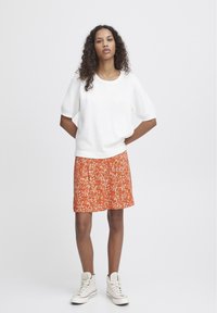 Witte korte mouw top gecombineerd met een oranje rok met luipaardpatroon. Draagt beige hoge sneakers en staat met de handen op de heupen.