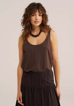 Žena sa kovrčavom smeđom kosom u bezručnom smeđem topu, crnom ogrlicom od perlica, crnoj suknji i malim tetovažama na lijevoj ruci na neutralnoj pozadini.