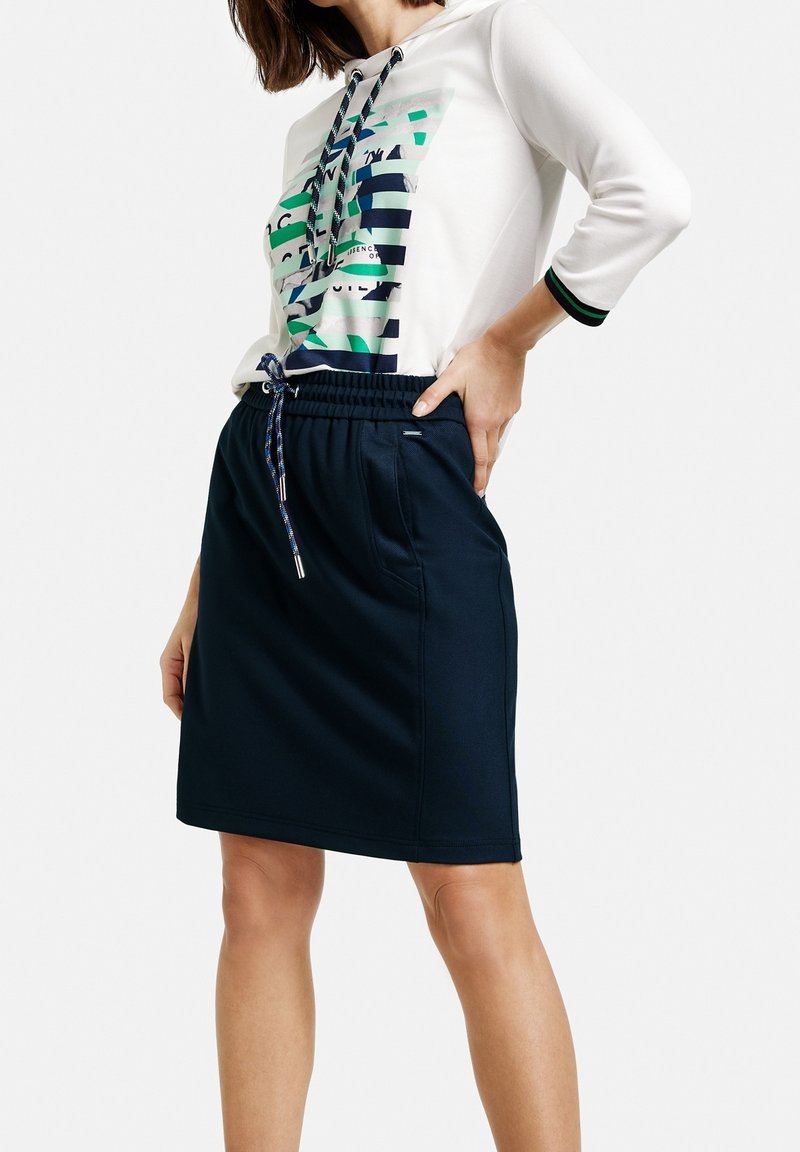 Femme portant un sweat à capuche blanc avec des rayures vertes et bleues et une jupe bleu marine à cordon, posant avec une main sur la hanche.