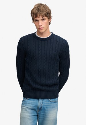 Jonge man met kort golvend haar, gekleed in een donkerblauwe kabelgebreide trui en lichtblauwe jeans tegen een effen witte achtergrond.