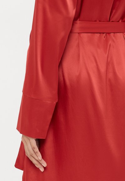Red satin garment with a smooth texture, featuring a belted waist and wide sleeves. Hand gently resting on the fabric.