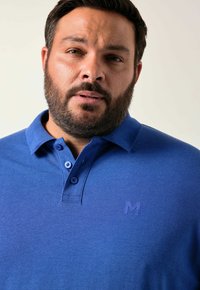 Sininen poolopaita, jossa on teksturoitu kangas, kolme nappia kauluksessa ja hillitty brodeerattu logo rinnassa.