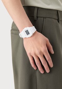 Montre numérique avec un bracelet en résine blanche, un cadran rectangulaire et un écran numérique gris affichant l'heure et les fonctions, portée à un poignet.