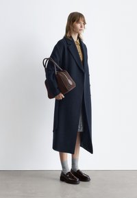 Langer navyblauer Wollmantel mit Revers, kombiniert mit einer braunen Wildledertasche. Das Model trägt graue Socken und dunkle Slipper. Der Hintergrund ist schlicht weiß.