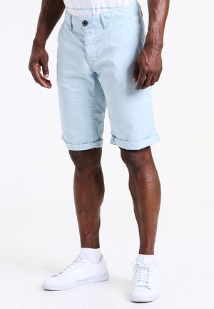 Homme portant un short bleu clair retroussé, des baskets blanches et des chaussettes blanches montantes, se tenant devant un fond blanc uni.