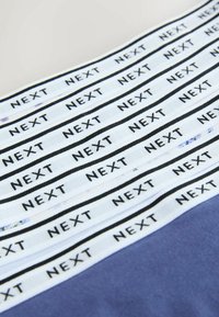 Weiße Stoffbänder mit dem Wort „NEXT“, mehrfach in Schwarz bedruckt, diagonal auf einer zweifarbigen Oberfläche angeordnet.