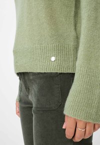 Maglione lavorato a maglia verde chiaro con orlo e polsini a coste. Un dettaglio di accento argento è visibile all'orlo inferiore. Abbinato a pantaloni in velluto a corda verde scuro.