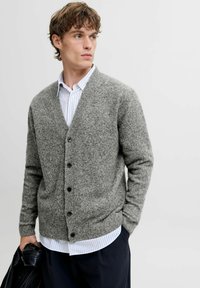 Grauer, gestrickter Cardigan mit V-Ausschnitt, langen Ärmeln, Knopfvrschluss und strukturiertem Finish, kombiniert mit einem weiß-blau gestreiften Hemd.