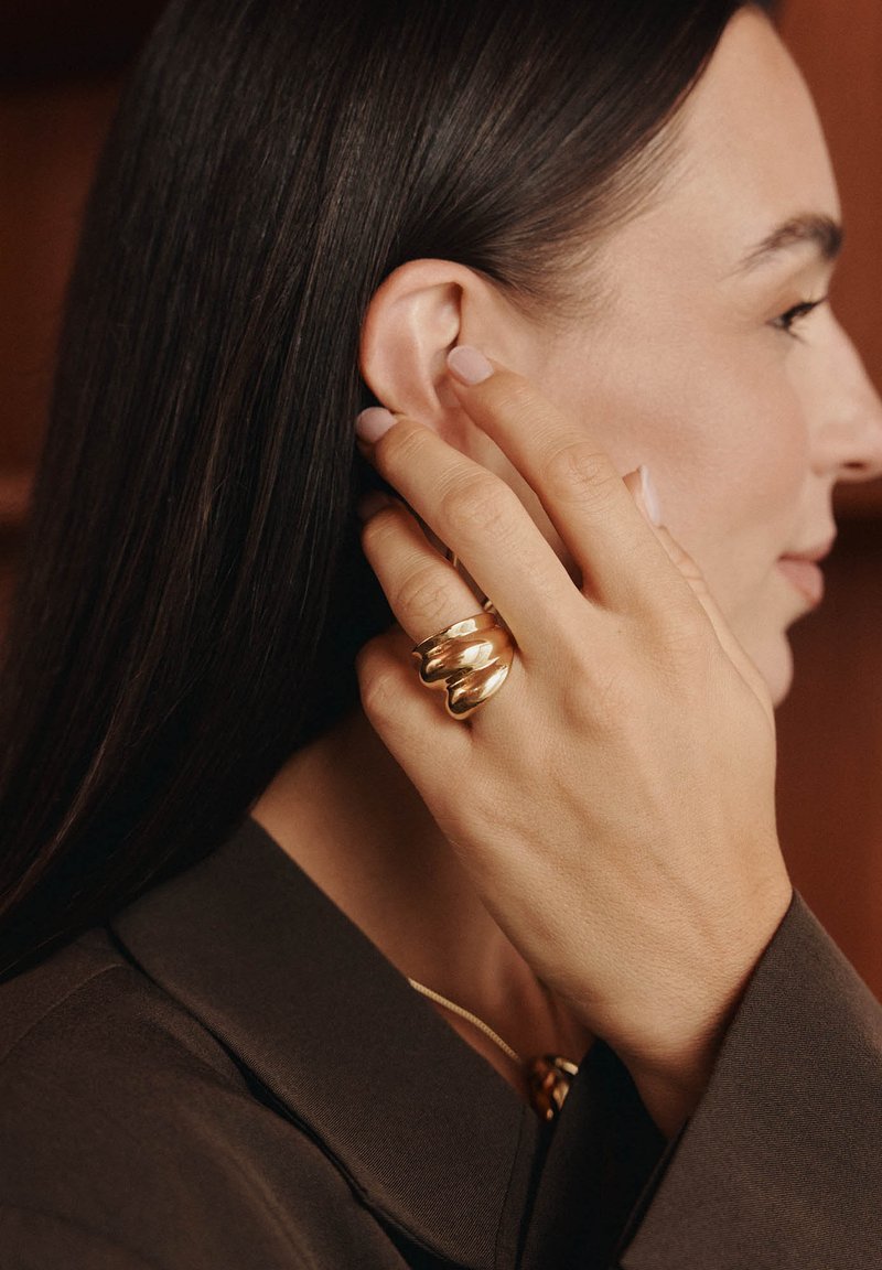 Goldene Spirale-Ring an einer Hand, glatte Oberfläche. Langes dunkles Haar, neutralfarbener Nagellack, trägt ein dunkles Shirt. Ohr teilweise sichtbar.