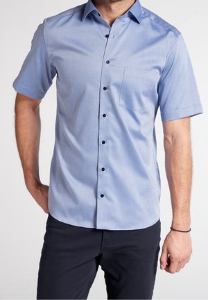 Uomo che indossa una camicia azzurra a maniche corte con taschino sul petto e pantaloni scuri, in piedi con una mano in tasca.