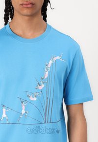 Gaiši zila kokvilnas T-krekls ar grafisko dizainu, kurā attēloti rozā un pelēki figūras, kas kāpj, un apakšā ir "adidas" logo.