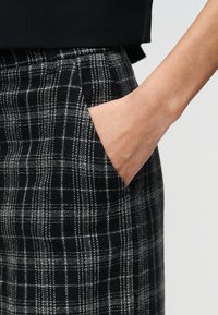 Χέρι μέσα στην εμπρόσθια τσέπη μαύρου και άσπρου καρό παντελονιού που φοριέται με μαύρο τοπ σε απλό φόντο.