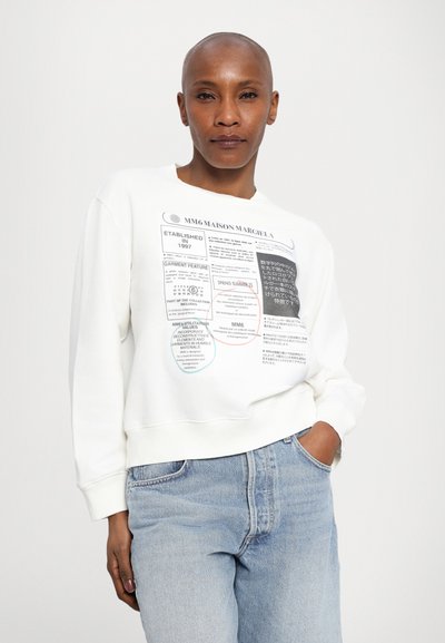 White sweatshirt featuring a printed label design with text in black, blue, and red. Ribbed cuffs and hem, relaxed fit. Worn with light blue jeans.