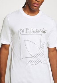 Vit bomullst-shirt med en svart omrissgrafik med texten "adidas" och en logotyp. Kortärmad med rund halsringning.