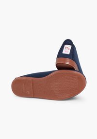 Scarpe in tela blu navale con una suola in gomma marrone texturizzata e un'etichetta che mostra un logo. Il design è privo di lacci e presenta una punta arrotondata.