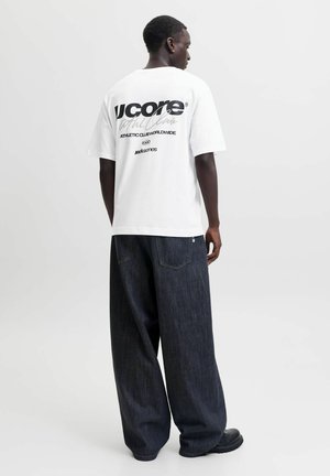 Mand står med ryggen til iført hvid t-shirt med grafik og løstsiddende mørkeblå jeans med sorte sko mod en ensfarvet lys baggrund.