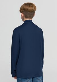 Granatowa koszula z długim rękawem i wysokim kołnierzem, wykonana z gładkiej, elastycznej tkaniny, o luźnym fasonie i minimalnej liczbie szwów.