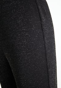 Fekete szövet leggings, diszkrét csillogással, sima textúrával és karcsú fazonnal. A részletek között szerepel egy oldalsó varrás a stílus érdekében.