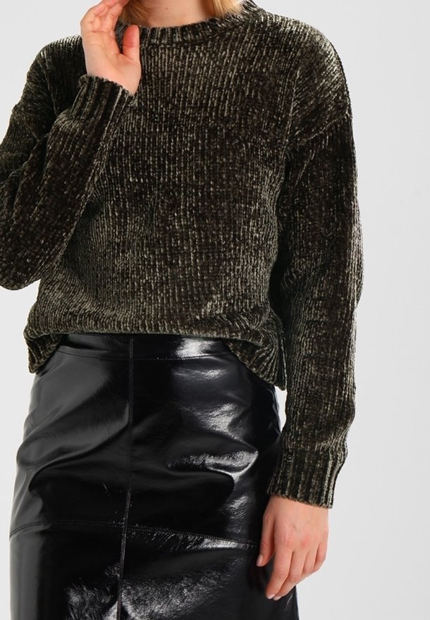 Femme portant un pull en velours côtelé vert foncé et une jupe en cuir noir brillante, debout avec une main près du visage et l'autre le long du corps.