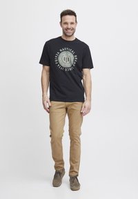 Sort bomulds t-shirt med et cirkulært grafik, der har "Nautical Brand Since 1924" i cream og grøn, kombineret med beige chinos og grå sko.