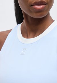 Κοντινό πλάνο ενός ατόμου που φοράει ανοιχτό μπλε αμάνικο τοπ με λευκή επένδυση και μικρό λευκό λογότυπο στο στήθος.