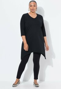 Zwarte tuniek met een V-hals, driekwartmouwen en een asymmetrische zoom, gedragen over zwarte leggings en gestreepte platte schoenen.