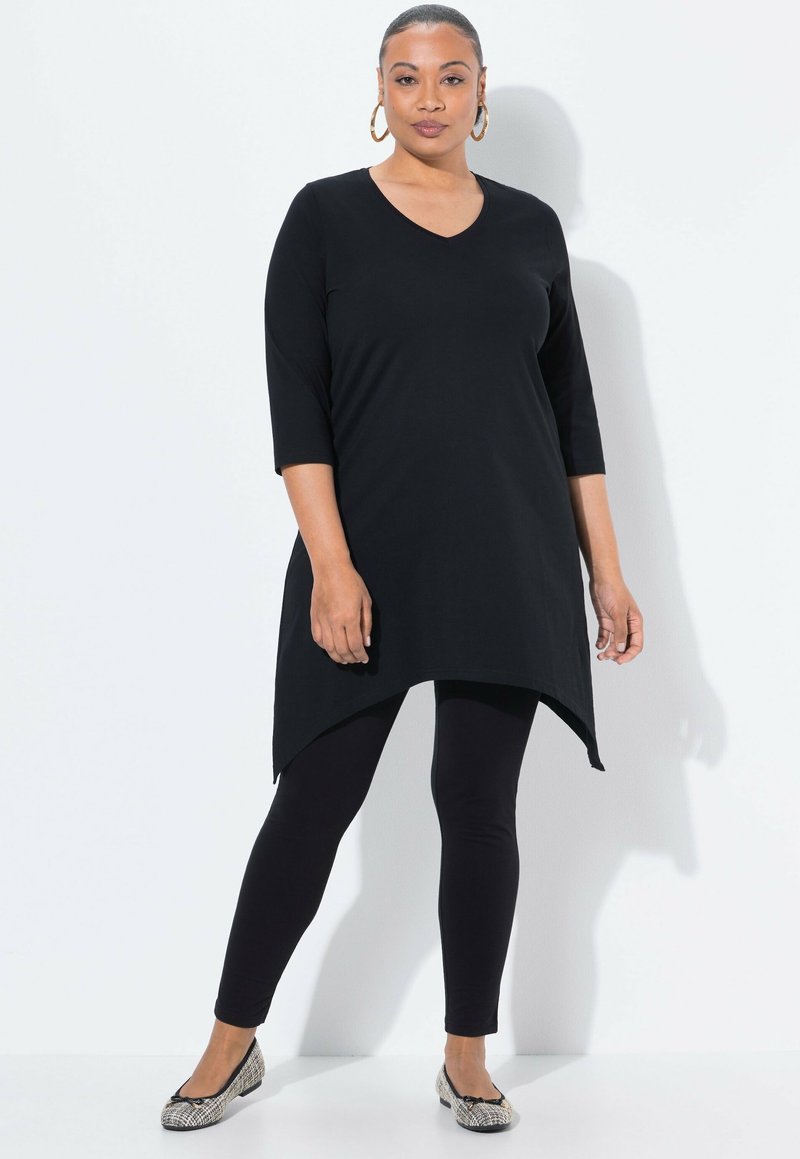 Zwarte tuniek met een V-hals, driekwartmouwen en een asymmetrische zoom, gedragen over zwarte leggings en gestreepte platte schoenen.