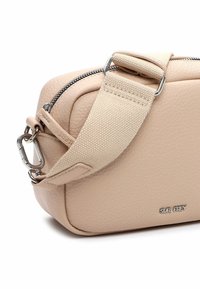 Bolso bandolera beige texturizado con correa ancha de lona ajustable, cremallera plateada, clip de metal y pequeño logo "SURI FREY" en la parte frontal.