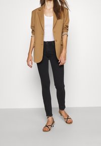 Ruskea räätälöity bleiseri valkoisen toppit paidan päällä, yhdistetty mustiin skinny-farkkuihin ja mustiin remmisandaaleihin. Liukas kangas ja jämäkkä muotoilu.