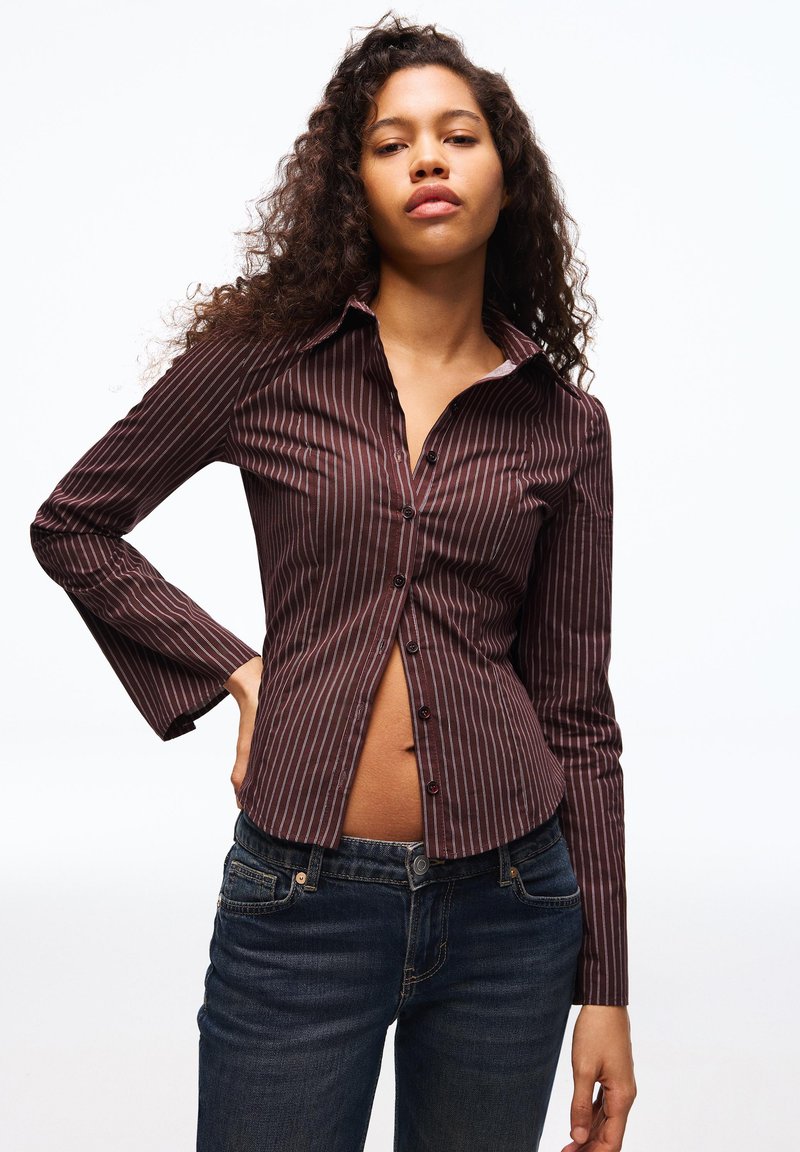 Brązowa koszula na guziki w paski z długimi rękawami, z ostro zakończonym kołnierzykiem i lekko rozszerzanymi mankietami, w połączeniu z ciemnoniebieskimi dżinsami.