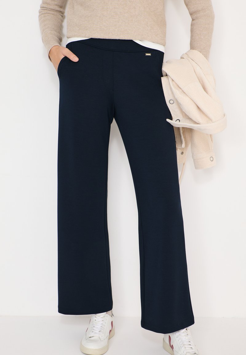 Personne portant un pantalon large bleu marine, un pull beige, des baskets blanches, tenant une veste beige boutonnée dans la main droite.