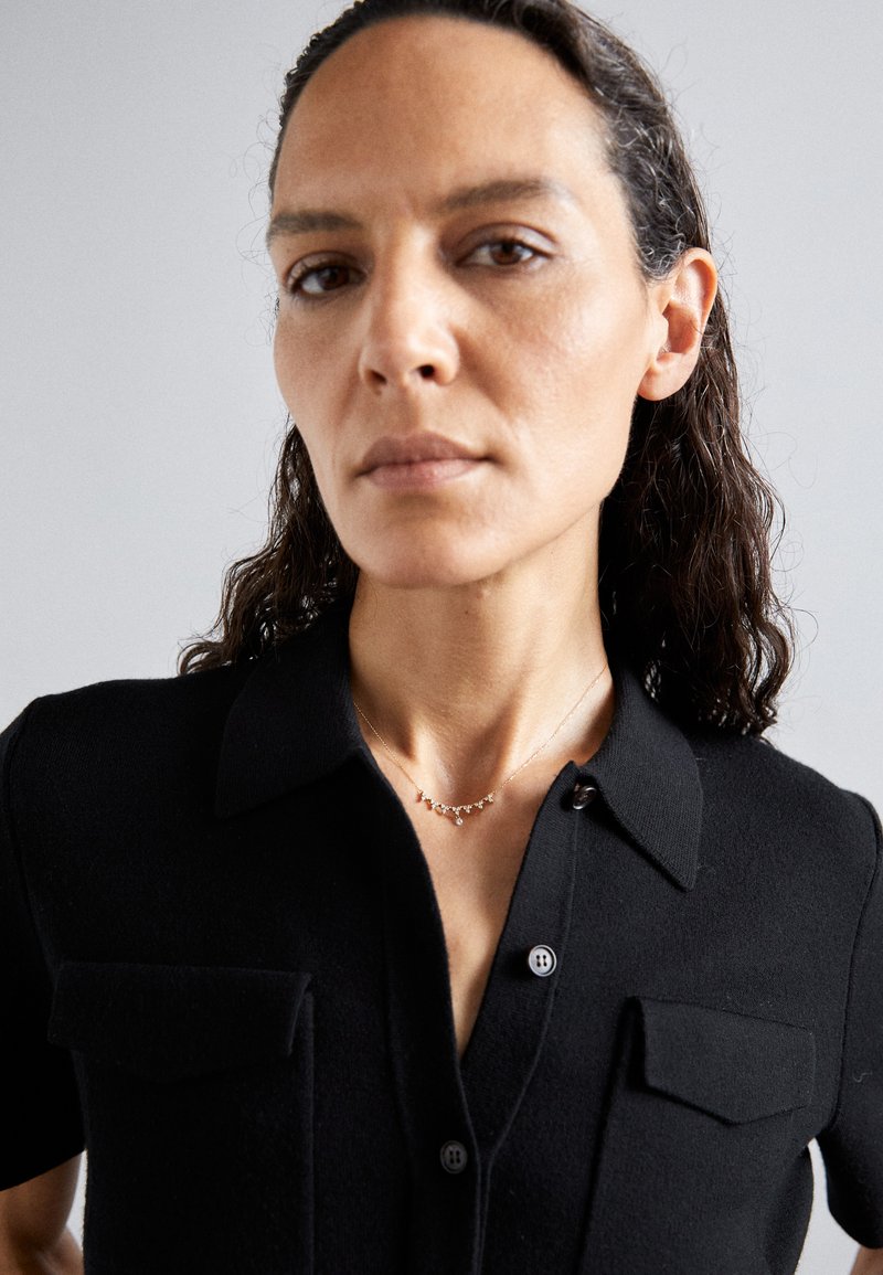 Camisa preta de manga curta com colarinho e dois bolsos frontais. Apresenta um colar delicado com pequenos acentos dourados e uma pedra preciosa clara.