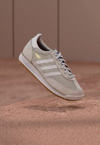 Bézs színű velúr sportcipő három fehér csíkkal, barna gumitalppal, fűzős kialakítással és arany logó díszítéssel az oldalon.