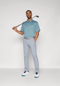 Lys blå pikéskjorte med korte ermer, lysegrå bukser, grått belte og hvite golfsko med blå detaljer, holder en golfkølle.