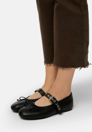 Black leather ballet flats with a round toe, bow detail, and an adjustable ankle strap featuring silver hardware. Texture is smooth.