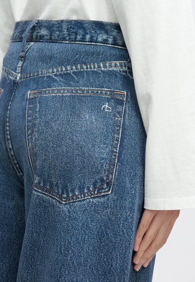 Niebieskie dżinsy z wysokim stanem, z widocznym przeszyciem, tylną kieszenią ozdobioną białym haftem oraz lekko zużytą fakturą.