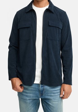 Mann trägt eine marineblaue Cordjacke mit Reißverschluss über weißem Hemd und hellblauen Jeans, steht mit den Händen nahe den Taschen.